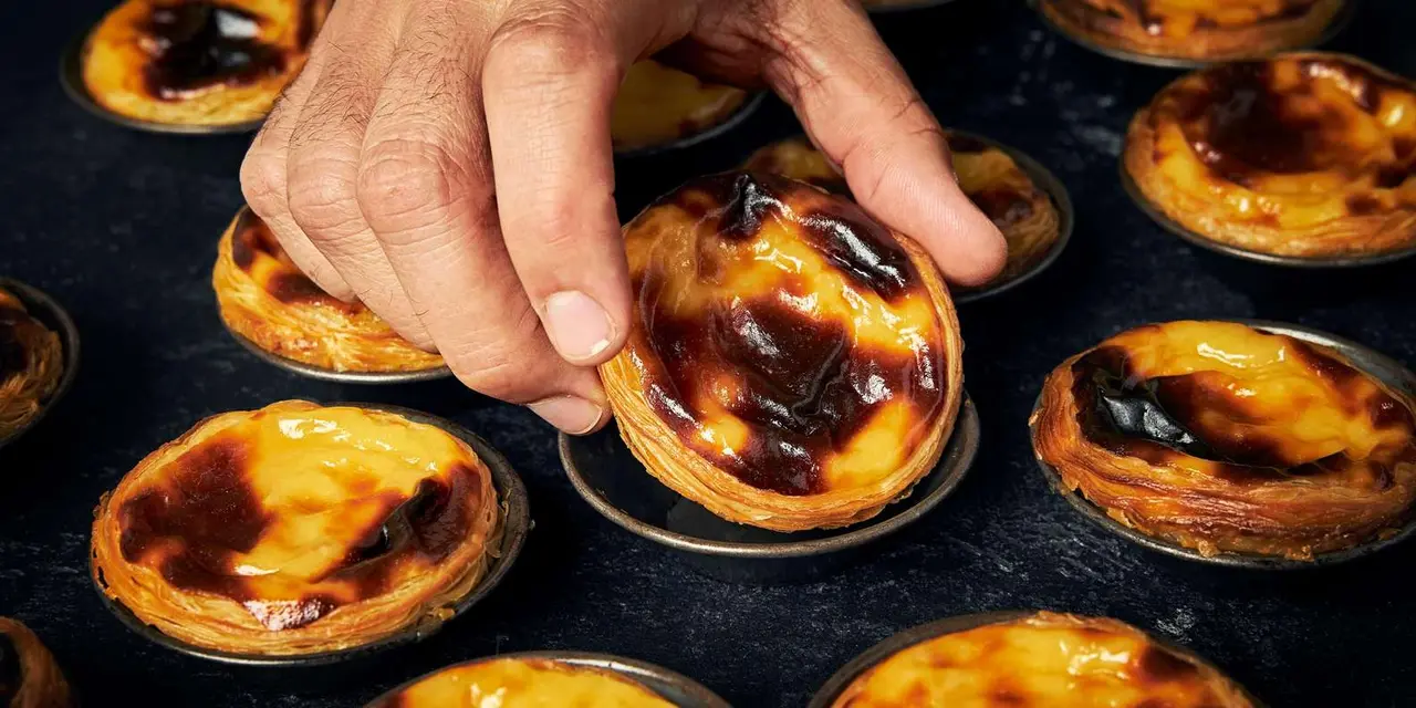 A hand lifting a freshly baked pastel de nata with a caramelised top from a tray of custard tarts