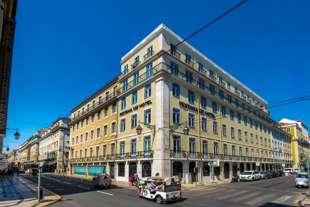 Pestana CR7 Lisboa's yellow corner building on Rua do Comércio in Baixa under a clear blue sky, with a tuk-tuk in the street