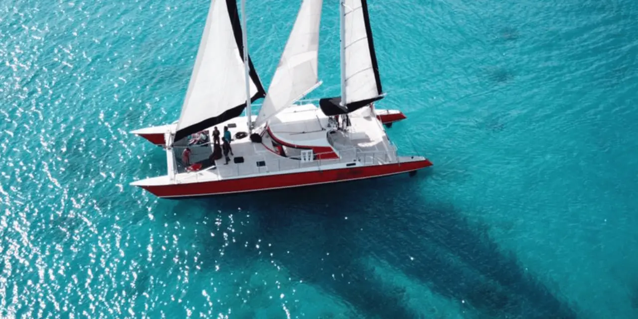 Aerial view of a white-sailed catamaran with a red hull on bright turquoise water