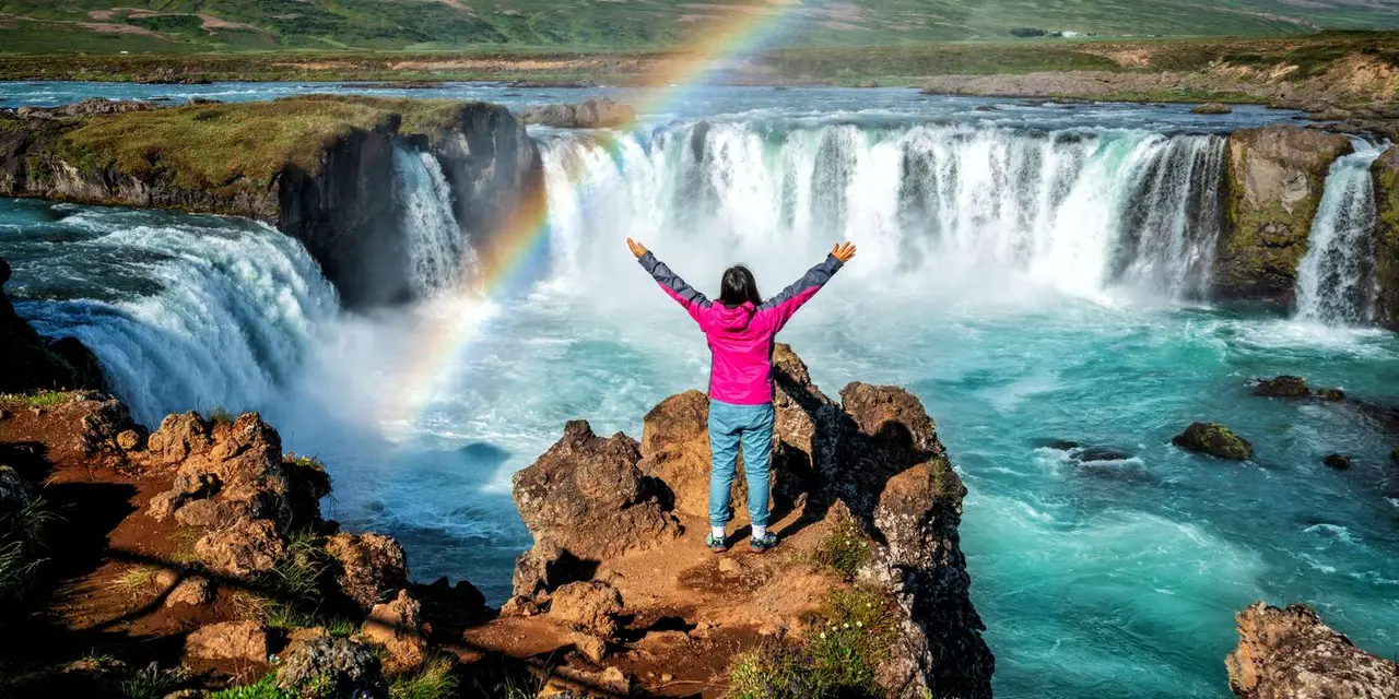 A person in a pink jacket standing on a rock with arms raised in front of Goðafoss with a rainbow over the spray