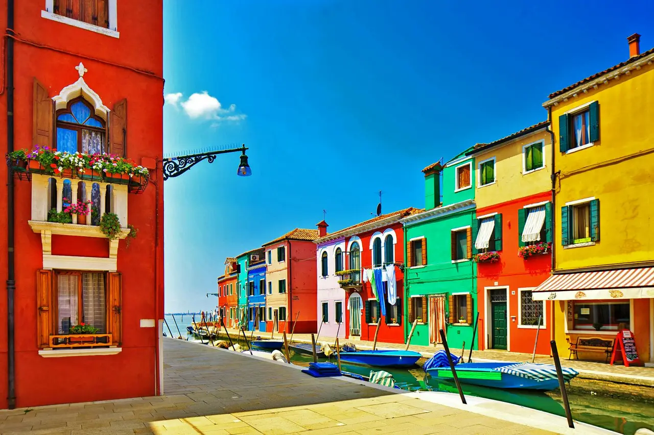 A street on Burano with red, pink, green and yellow houses along a canal under a blue sky