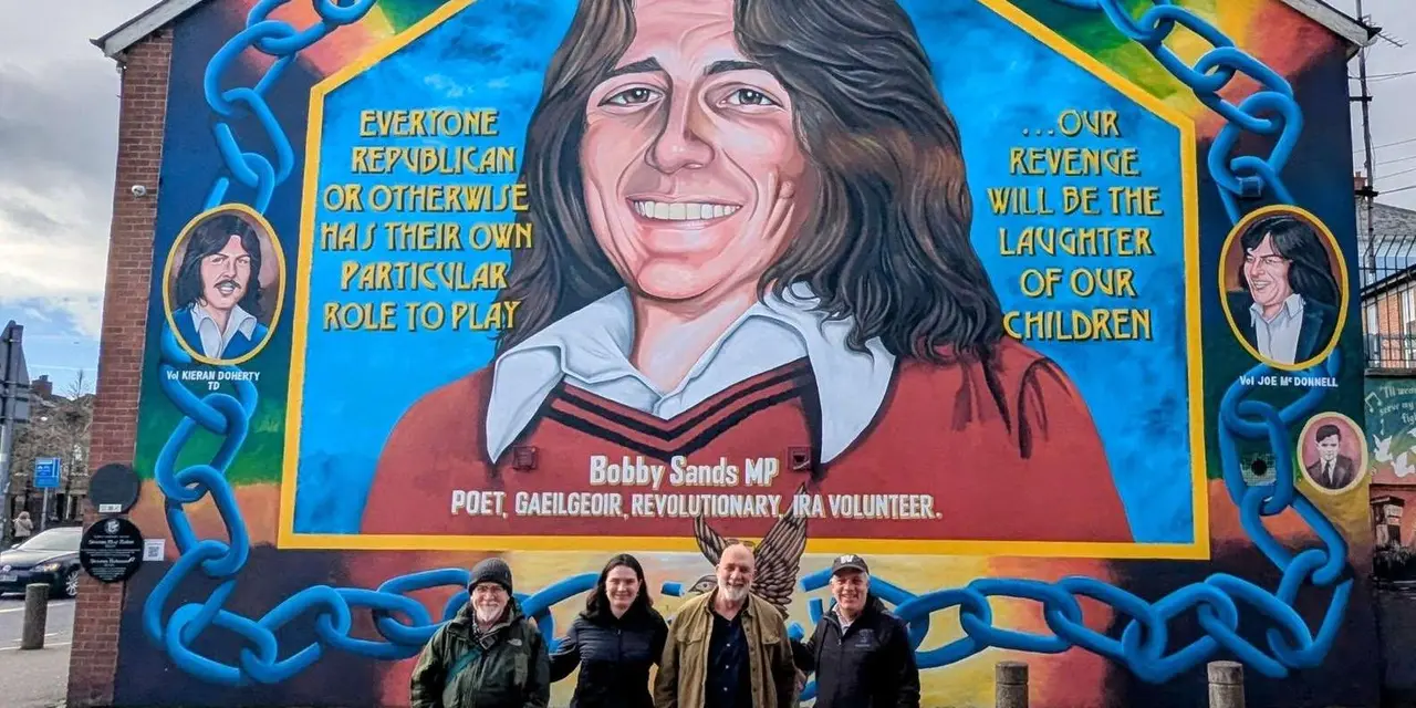 A large chain-bordered mural of a smiling man in a red jumper on a gable wall, with four visitors standing beneath it