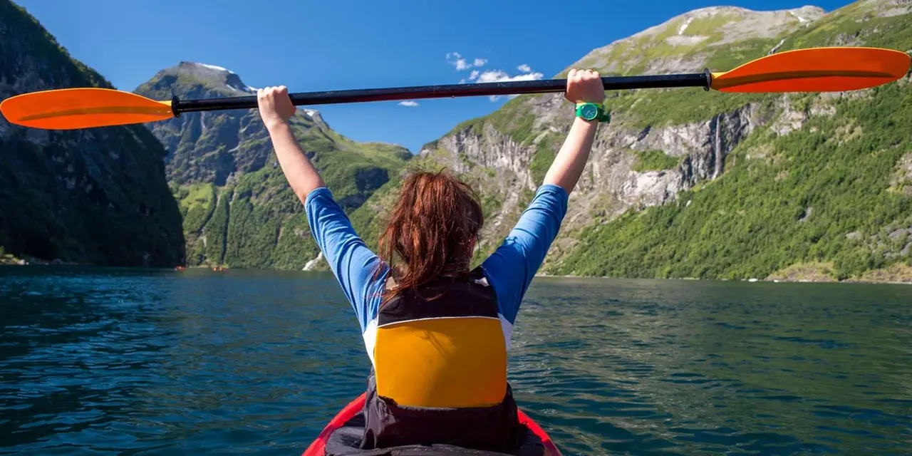 A paddler raising a paddle overhead from the bow of a red kayak on the fjord