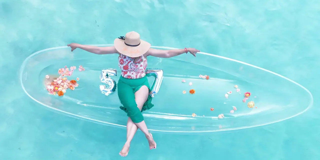 A woman in a sun hat and green skirt sitting in a clear kayak decorated with flowers, floating on pale turquoise water