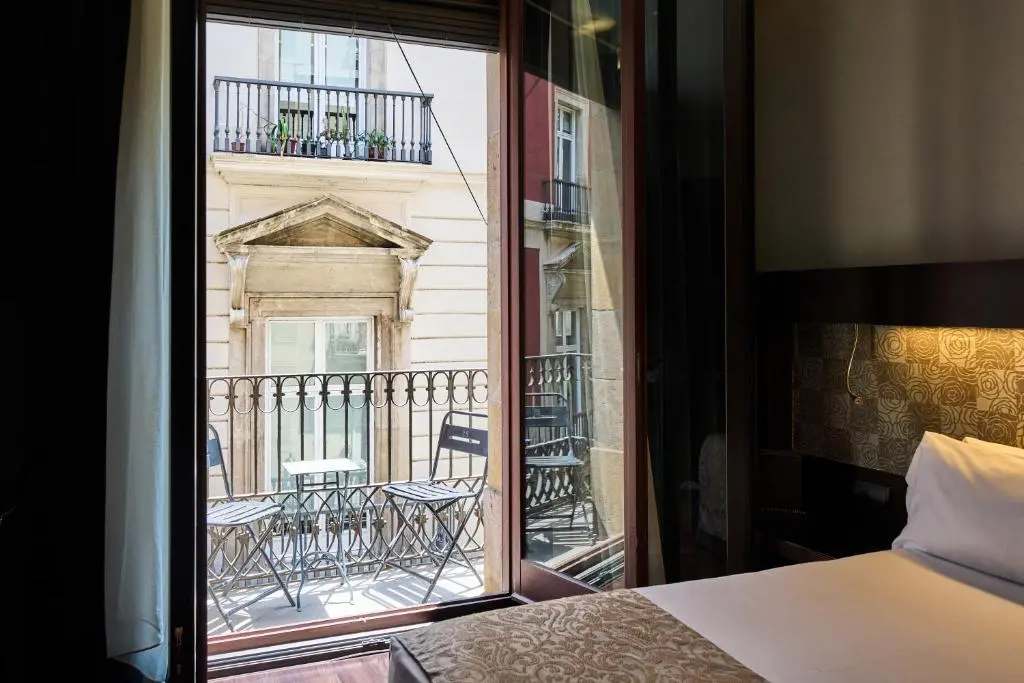 A Catalonia Port guest room with its door open onto a small balcony with a bistro table, facing an old stone facade across Carrer Ample