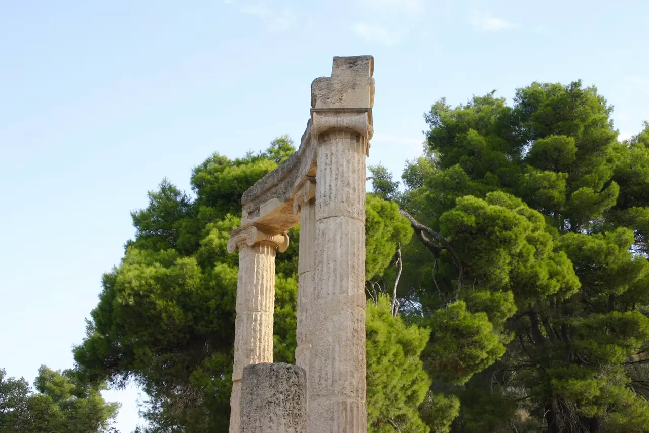 Two weathered stone columns rising against a pale sky beside green pine trees at Ancient Olympia, about 34 km inland from Katakolon