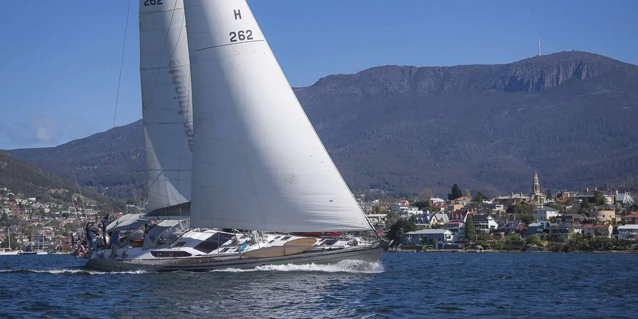White sails of a yacht heeling on blue water with the hills behind Hobart