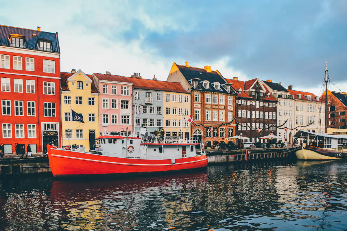 Red and yellow Nyhavn townhouses along the canal in Copenhagen, with a bright red boat moored in front and a tall ship's mast at the right