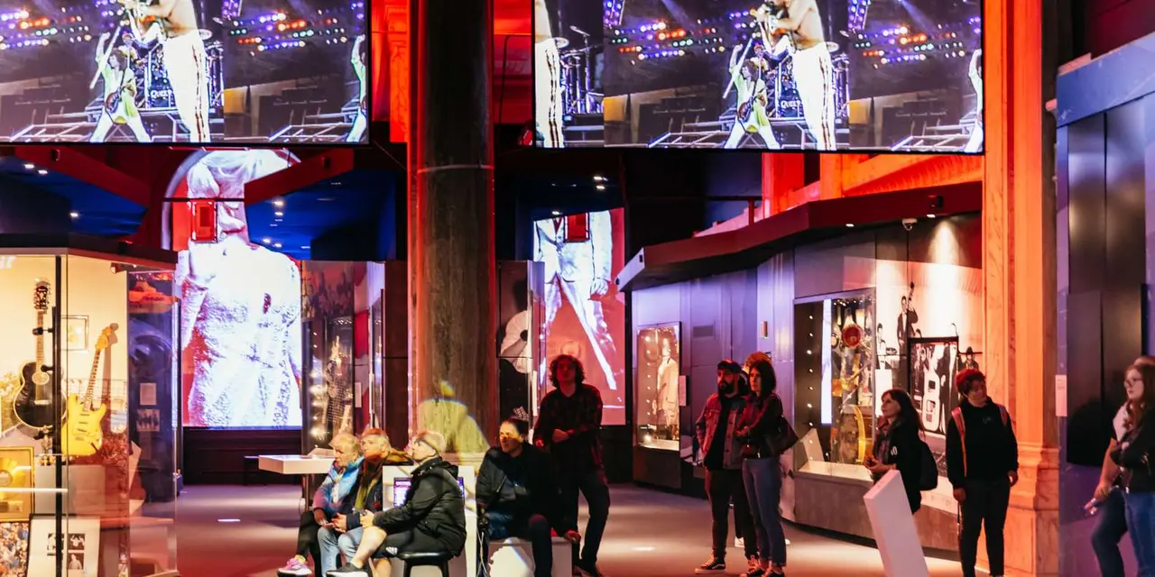 Visitors in a dim exhibition hall lit by large video screens showing a Queen concert, with glass cases of instruments beside them