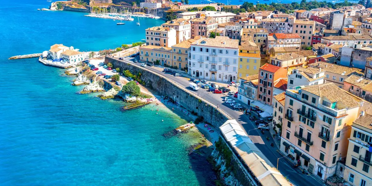 Aerial view of Corfu Town's pastel seafront buildings above turquoise water, with the Old Fortress and the sea behind