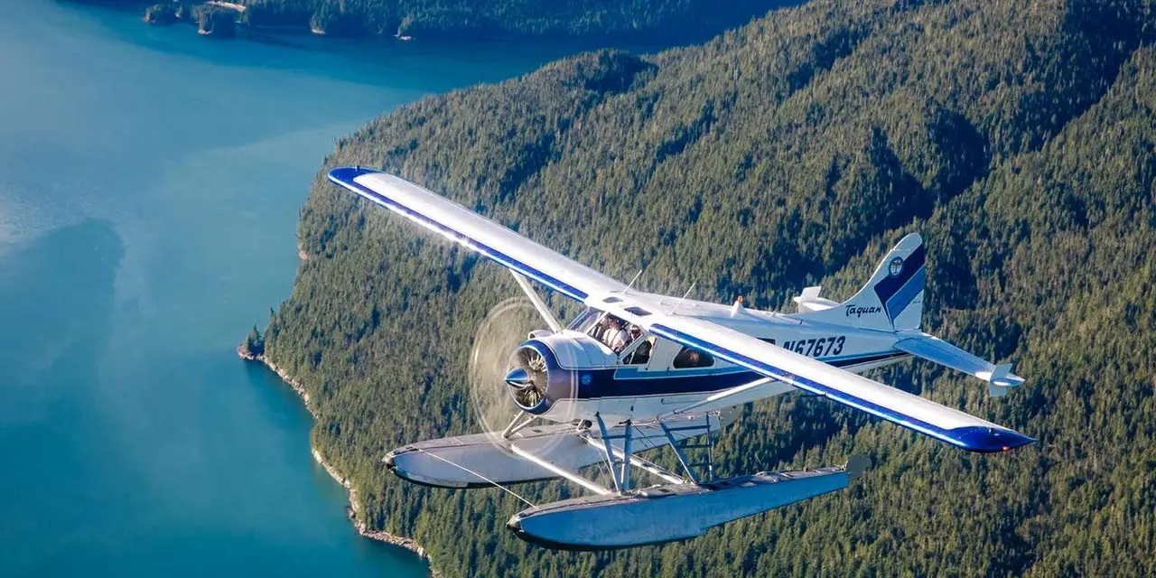 A blue-and-white floatplane flying low over forested hills and a winding inlet