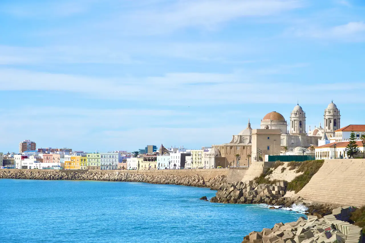Cádiz seen across a bright blue bay, with the golden-domed cathedral and its two bell towers rising from the white old town on the right and open sky to the left