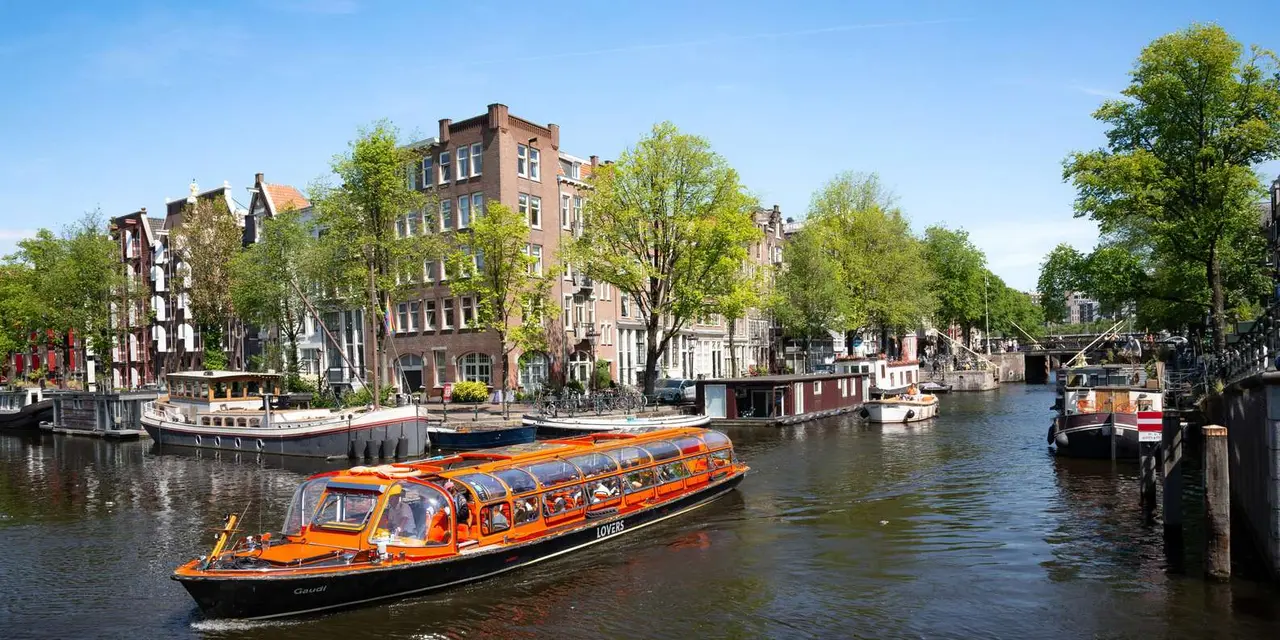 An orange glass-roofed canal boat passing moored houseboats and brick canal houses under a bright blue sky
