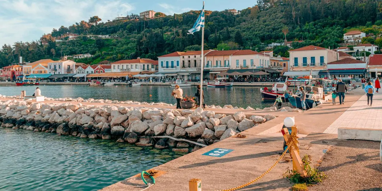 Katakolon's harbour waterfront with a Greek flag, a breakwater and pastel buildings below green hills