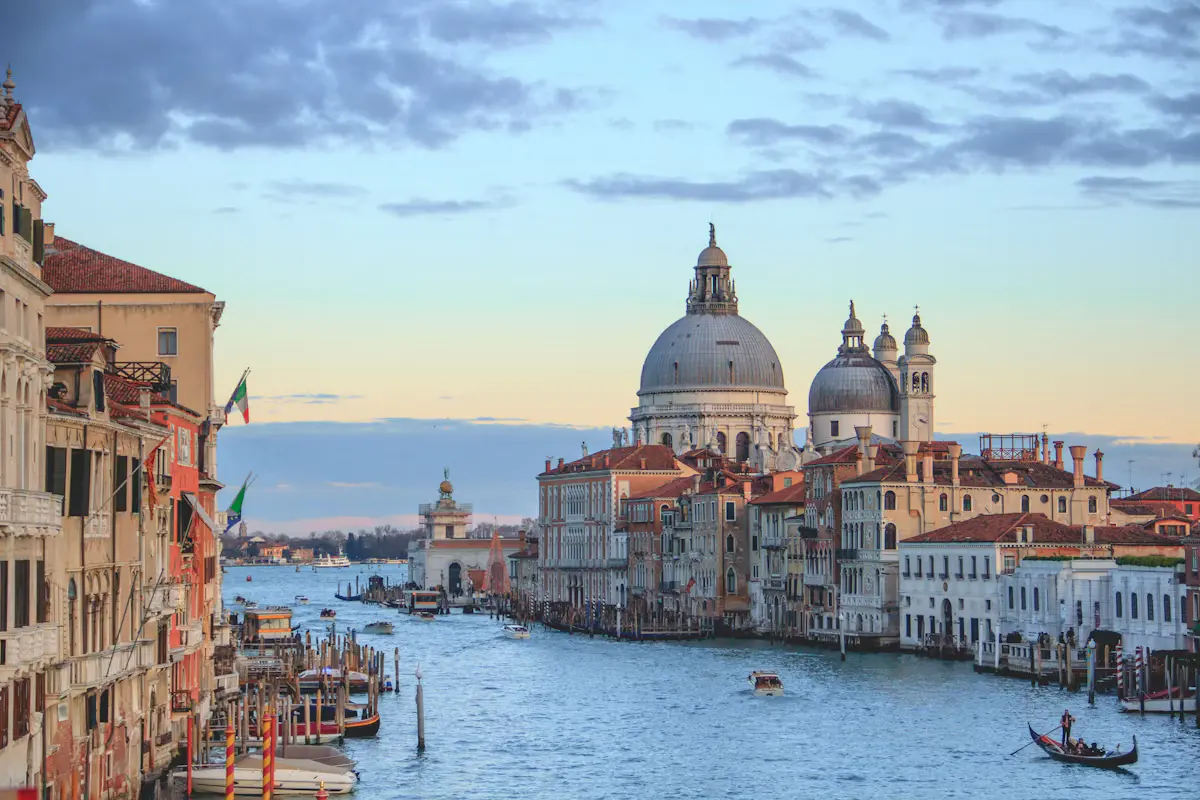 The Grand Canal in Venice seen from the Accademia Bridge, with the white dome of Santa Maria della Salute, palazzi along the water and a gondola in the foreground under a soft blue sky
