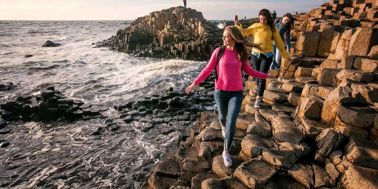 Three visitors stepping across the Giant's Causeway's hexagonal basalt columns beside breaking waves in low evening light