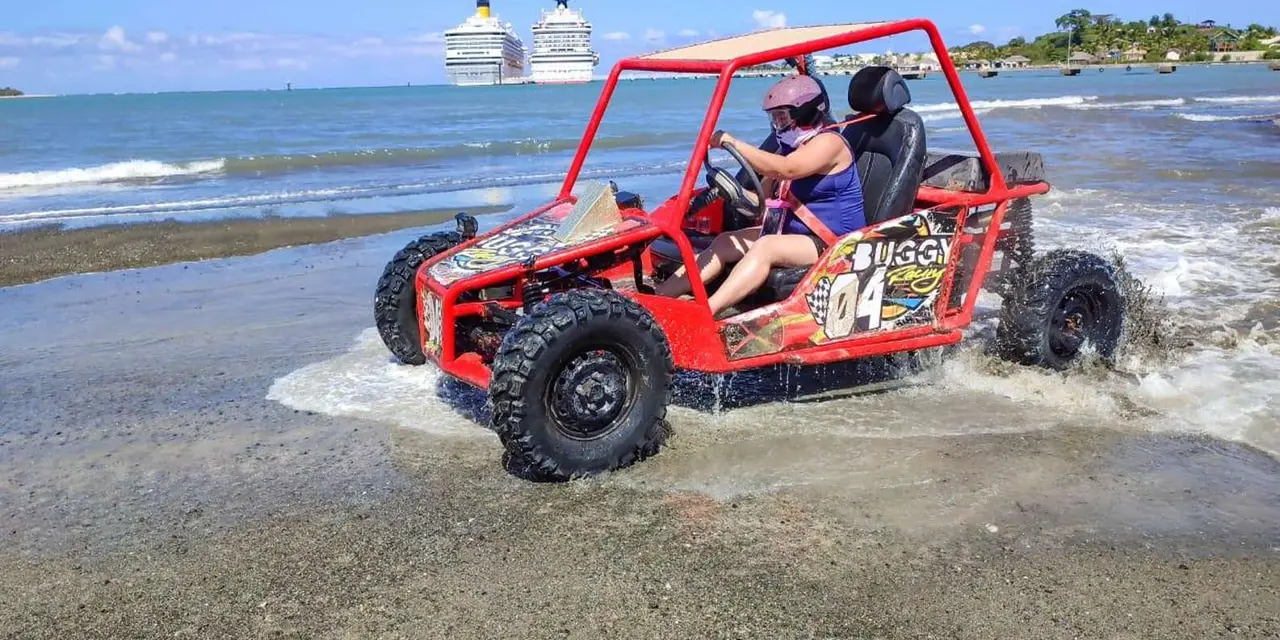 A red two-seat buggy splashing through shallow water on a beach, with cruise ships docked in the background