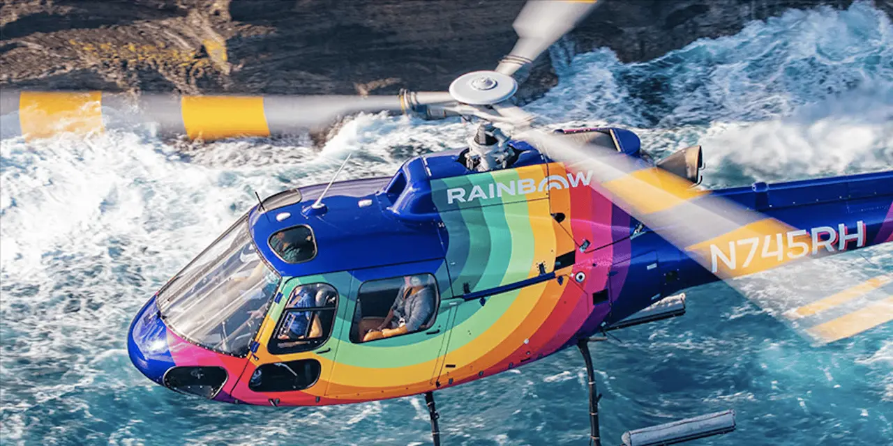 A rainbow-striped helicopter flying low over blue ocean beside a rocky coast