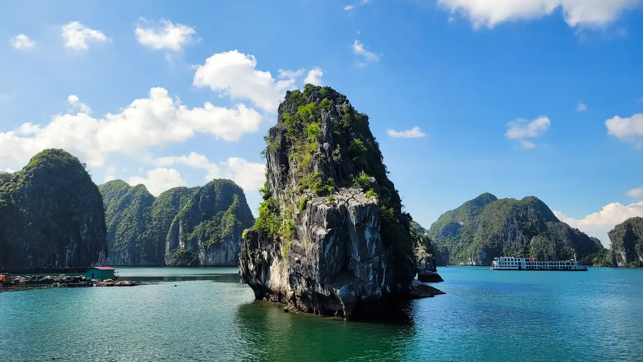 A tall rock stack rising from turquoise water in Ha Long Bay, with forested limestone islands and a white tour boat under a bright blue sky with scattered clouds