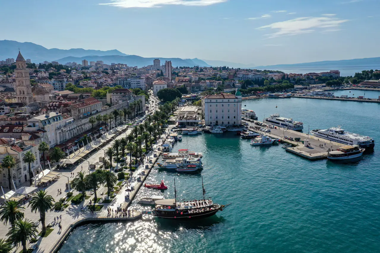 Split's palm-lined Riva promenade and harbour from above, with the cathedral bell tower, moored boats and ferries in turquoise water under a blue sky