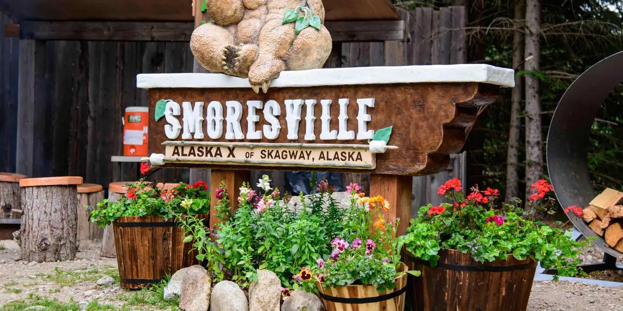 A carved wooden Smoresville sign among flower barrels at a camp near Skagway