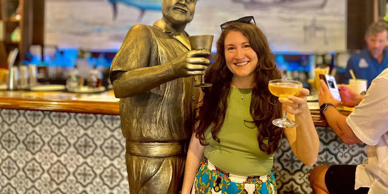 A woman smiling beside a bronze statue of Hemingway holding a cocktail glass, at a tiled bar