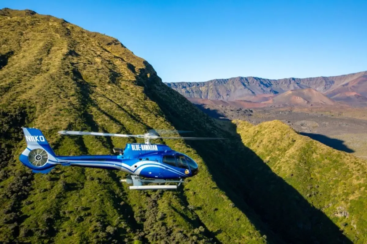 A blue helicopter flying along a green ridge with a reddish volcanic crater in the background