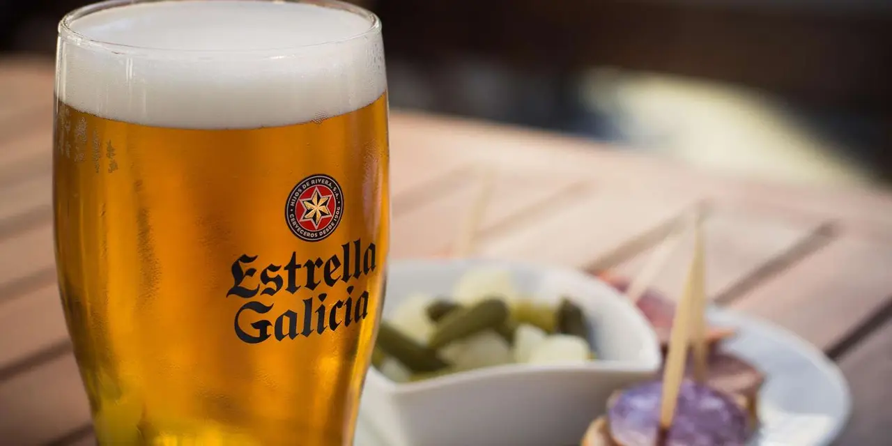 A glass of Estrella Galicia beer next to small dishes of tapas on a wooden table