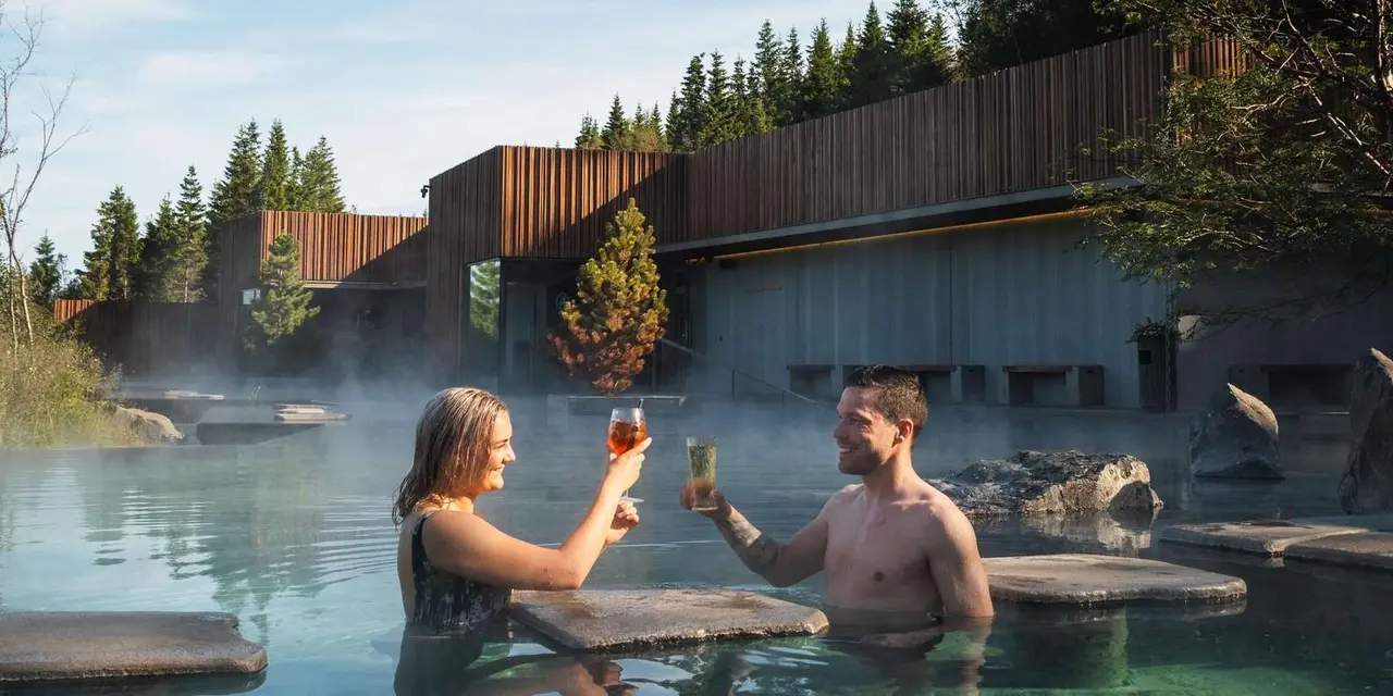 Two people toasting with drinks in a steaming geothermal pool, with a wooden spa building and pine trees behind