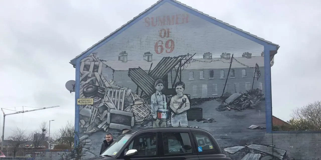 A black taxi parked in front of a grey gable-end mural of two boys and a pile of rubble, with the words Summer of 69 above them