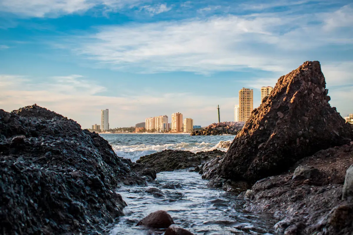 Waves washing between dark rocks on the Mazatlán shore, with a row of tall seafront buildings in the distance under a pale sky with streaks of cloud