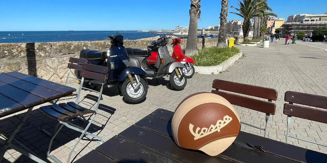 Scooters parked on a palm-lined seafront promenade, with a Vespa helmet resting on a bench in the foreground