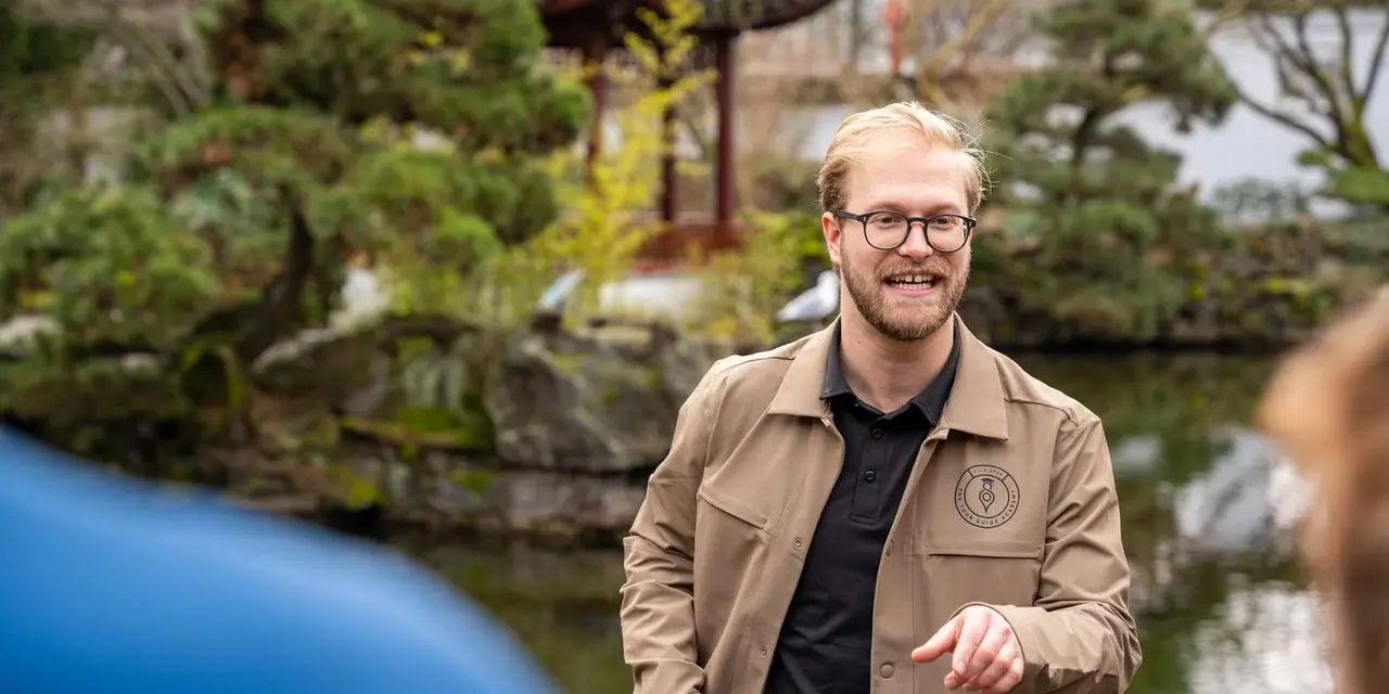 A smiling guide in glasses and a tan jacket talking to a group in a garden with a pond and pine trees