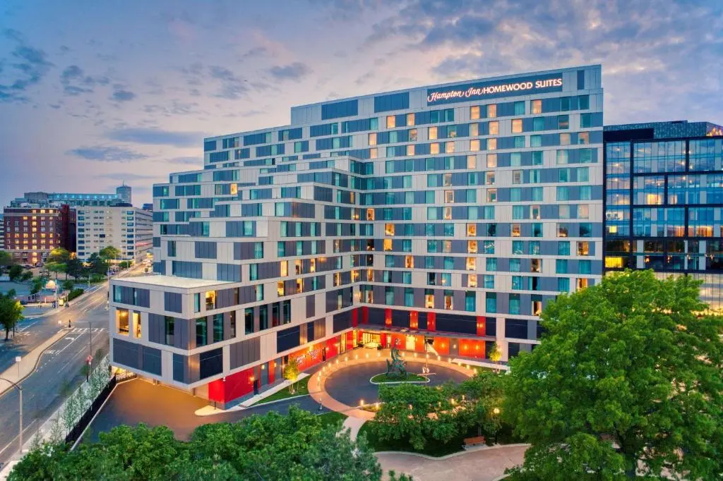 The Hampton Inn and Homewood Suites building in Boston's Seaport at dusk, its lit windows above a round driveway and trees
