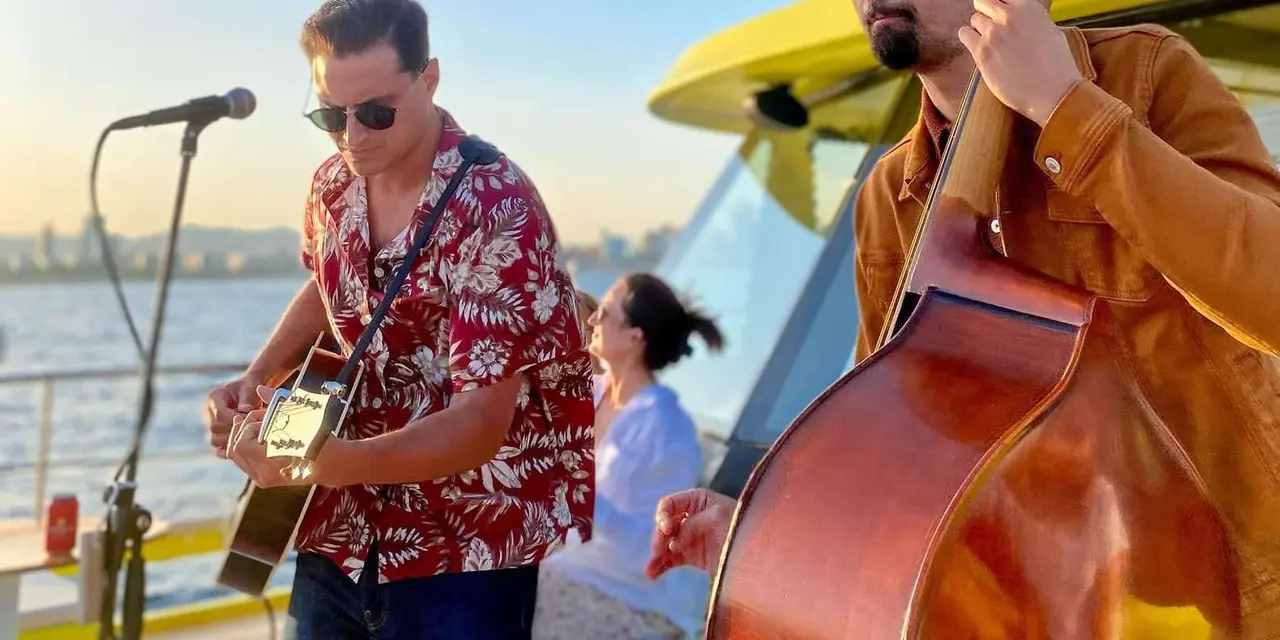 A guitarist in a red floral shirt and a double-bass player performing on the deck of a yellow-trimmed catamaran, with the sea and city skyline behind