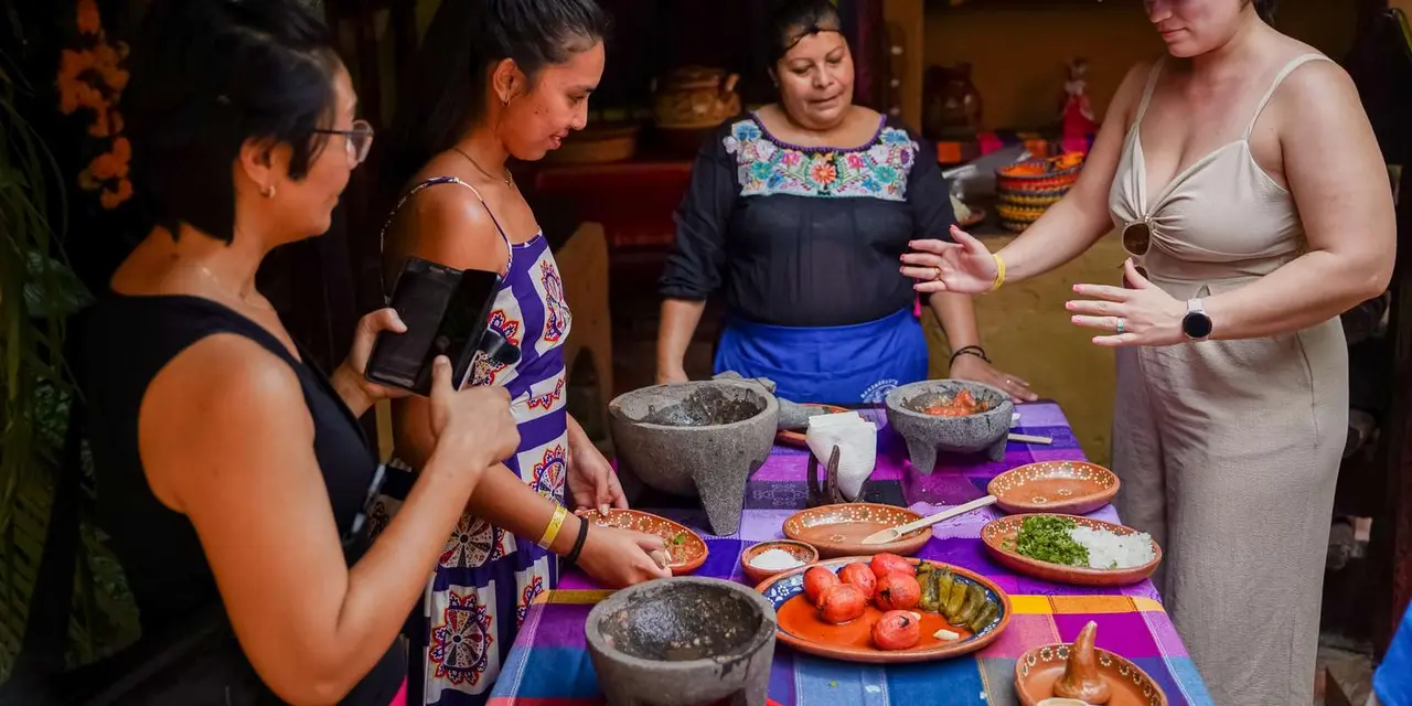 Women in aprons around a long table of stone molcajete bowls, tomatoes and chiles, with a cook in an embroidered blouse