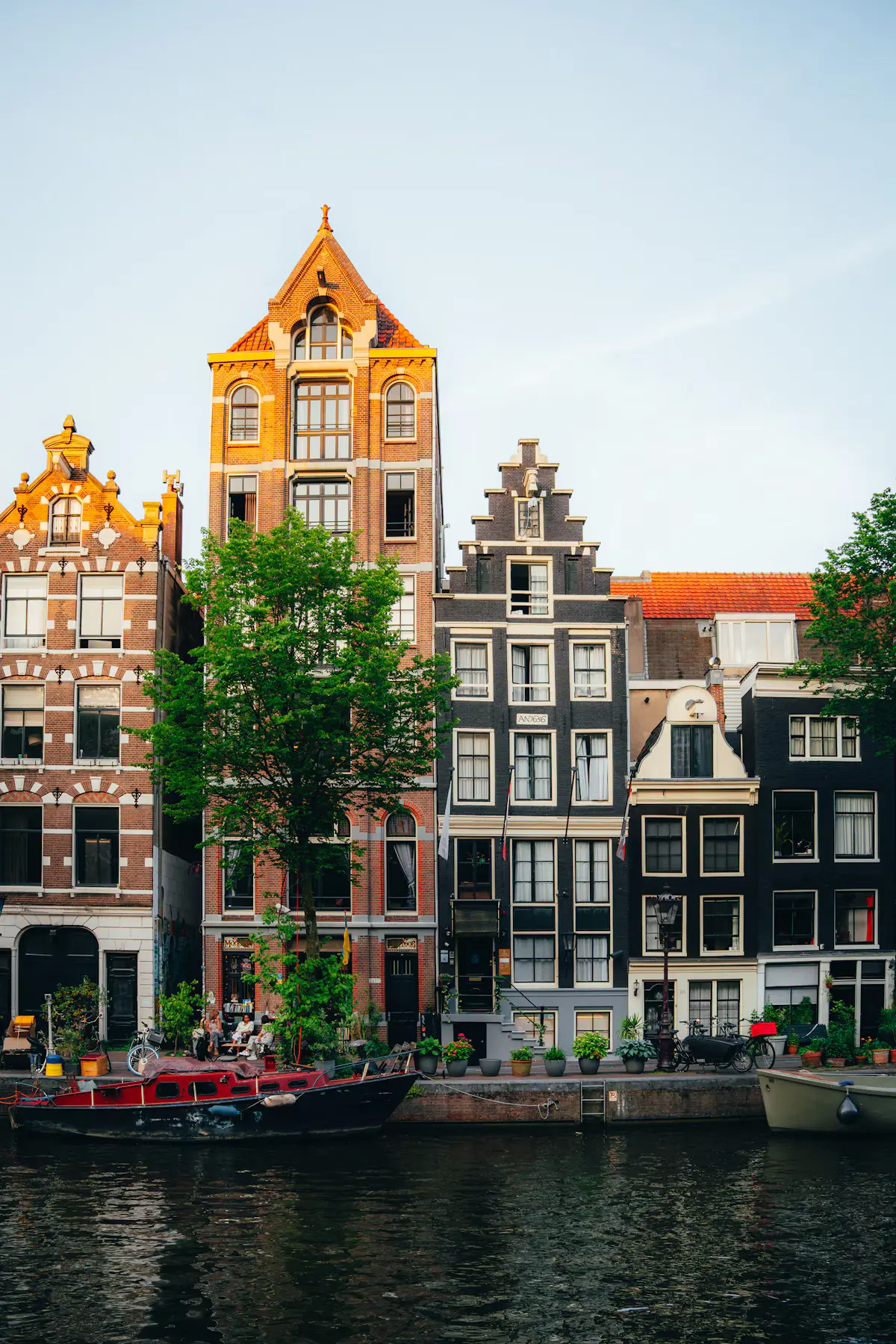 Row of tall brick and black-fronted Dutch canal houses in warm evening light, with a red boat moored at the canal's edge in Amsterdam