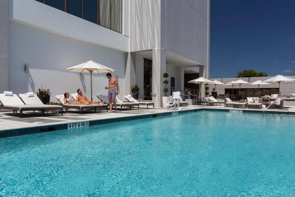 Guests on loungers beside EB Hotel Miami's turquoise rooftop pool with white umbrellas under a clear sky