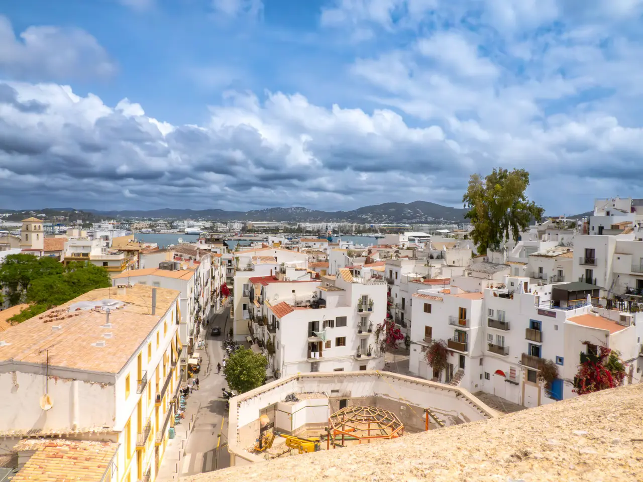 Whitewashed houses and a narrow street in Ibiza Town seen from the old town walls, with the harbour and hills in the distance under a cloudy sky