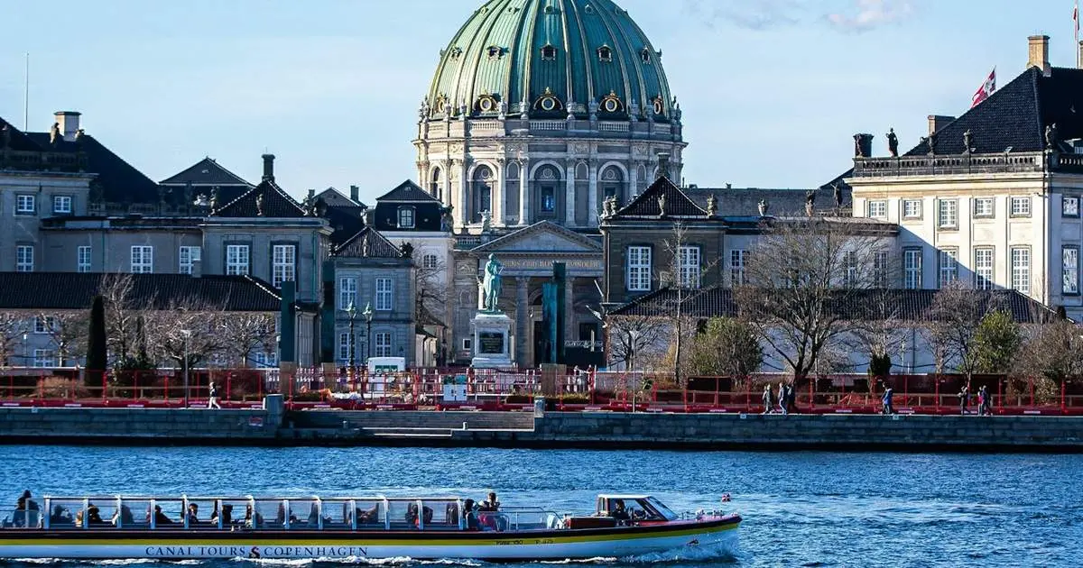 A canal tour boat with passengers crossing the harbor in front of a green copper-domed church and neoclassical palace buildings