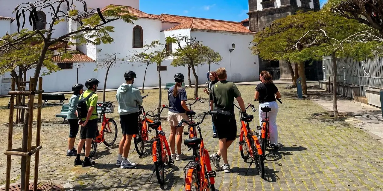 A group of cyclists with orange bikes standing on a cobbled square beside a whitewashed church while their guide talks