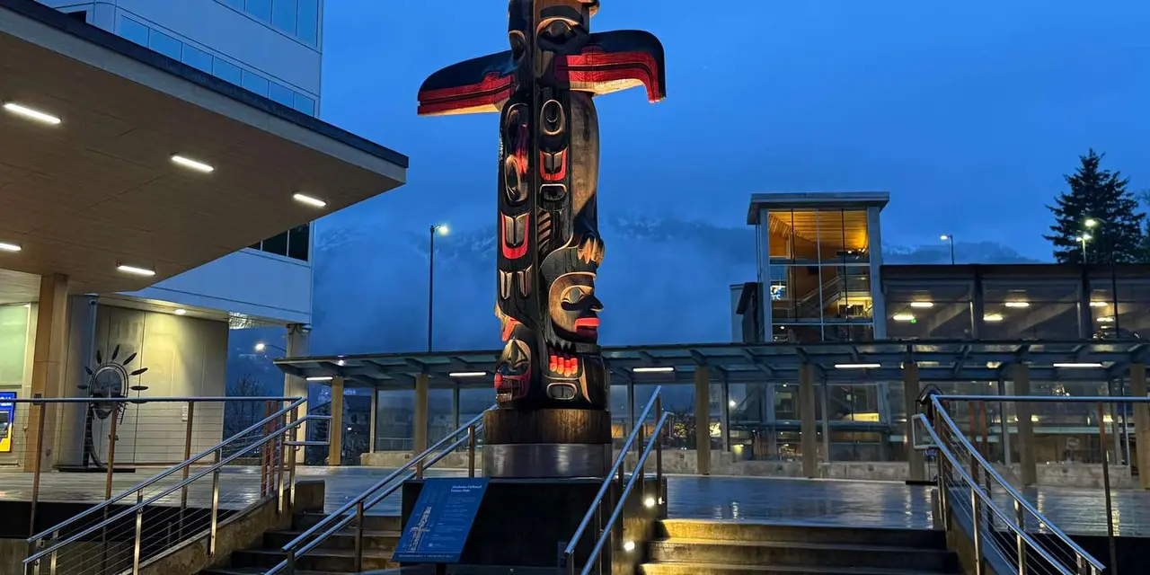 A carved totem pole with red and black outstretched eagle wings, lit at dusk in front of a glass-fronted building with mist over the mountain behind
