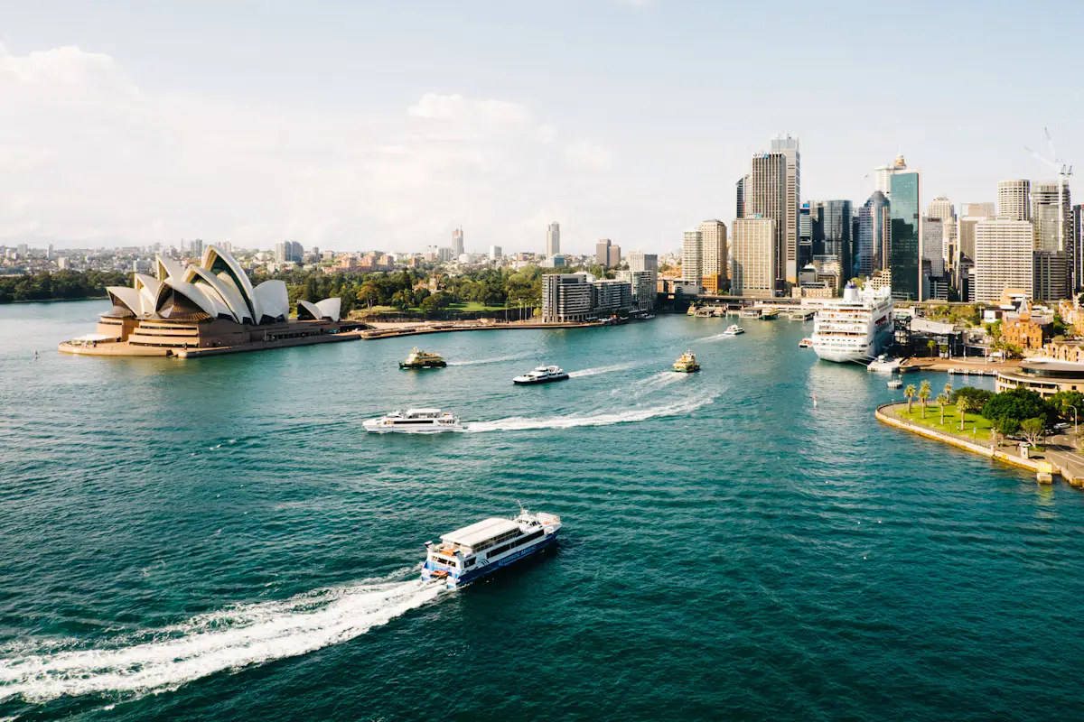 Sydney Harbour on a sunny day from above, with the Opera House on the left, ferries crossing Circular Quay and a white cruise ship docked at the Overseas Passenger Terminal below the city skyline