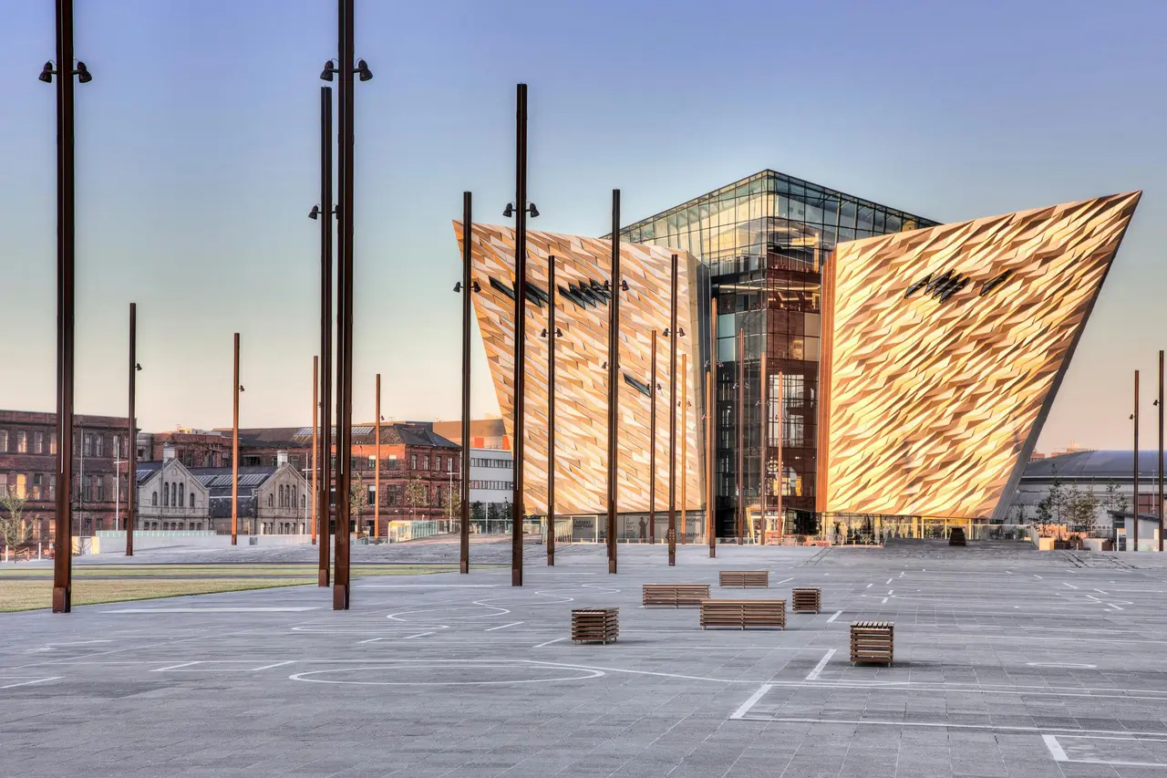 Titanic Belfast's angular gold-and-glass facade on an empty paved plaza at dusk, with rust-coloured lamp posts in the foreground