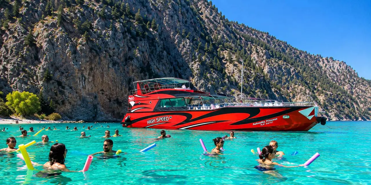 A red speedboat moored in turquoise water below a rocky cliff, with swimmers in the water around it