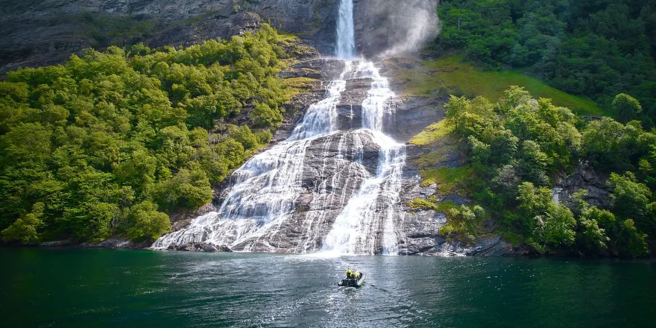 A tall waterfall cascading down a cliff into the fjord, with a small boat at its foot