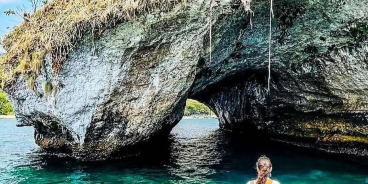 A natural rock archway over green water at Los Arcos, with a person in a sun hat in the water at the bottom right