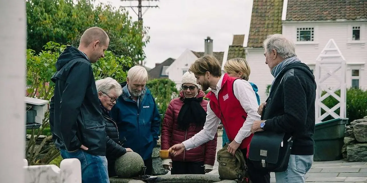 A guide in a red vest showing a small group an object beside a white wooden house