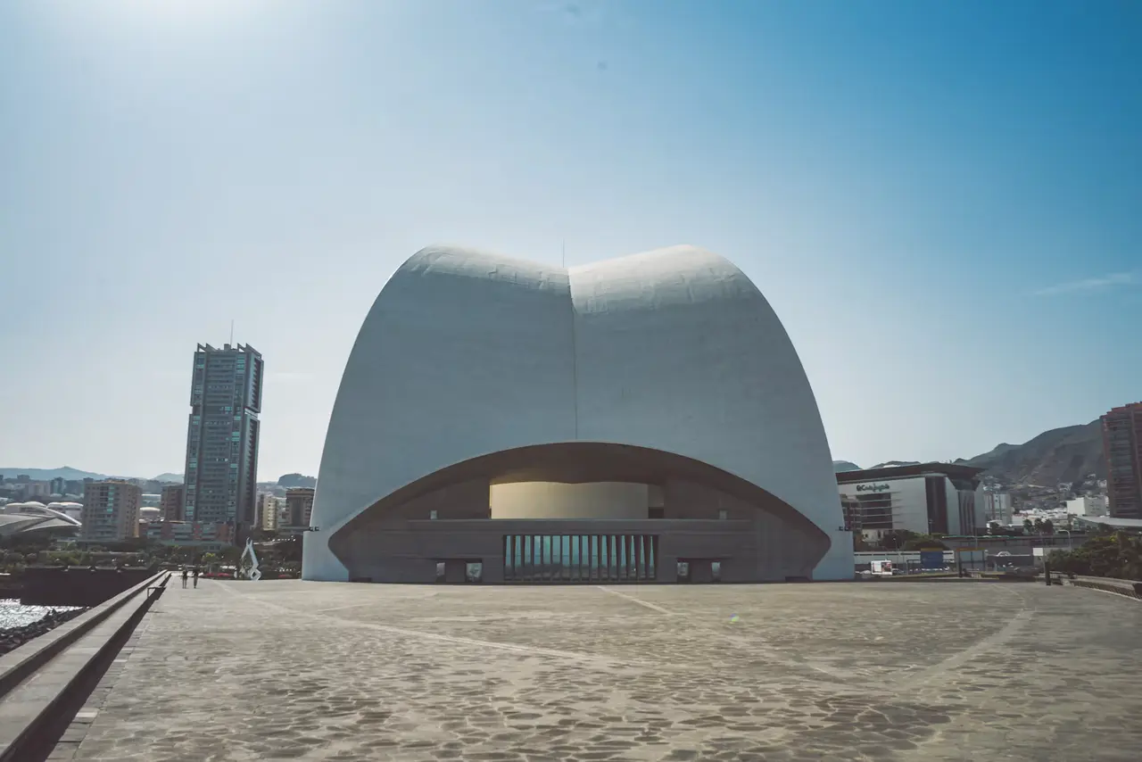 The white curved concrete shell of the Auditorio de Tenerife rising above a stone plaza, with apartment towers and mountains behind it under a clear blue sky