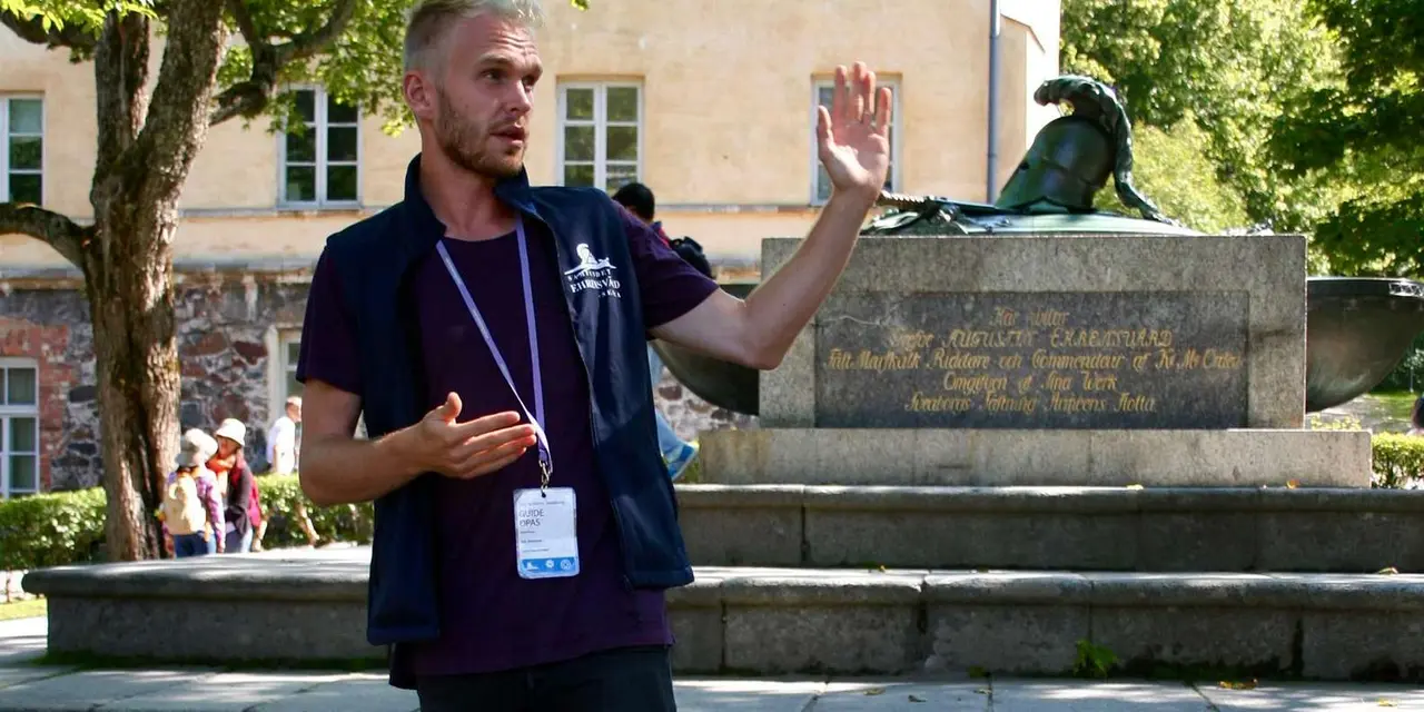 A guide in a navy vest and lanyard gestures beside a stone monument and green trees, with an old yellow building behind
